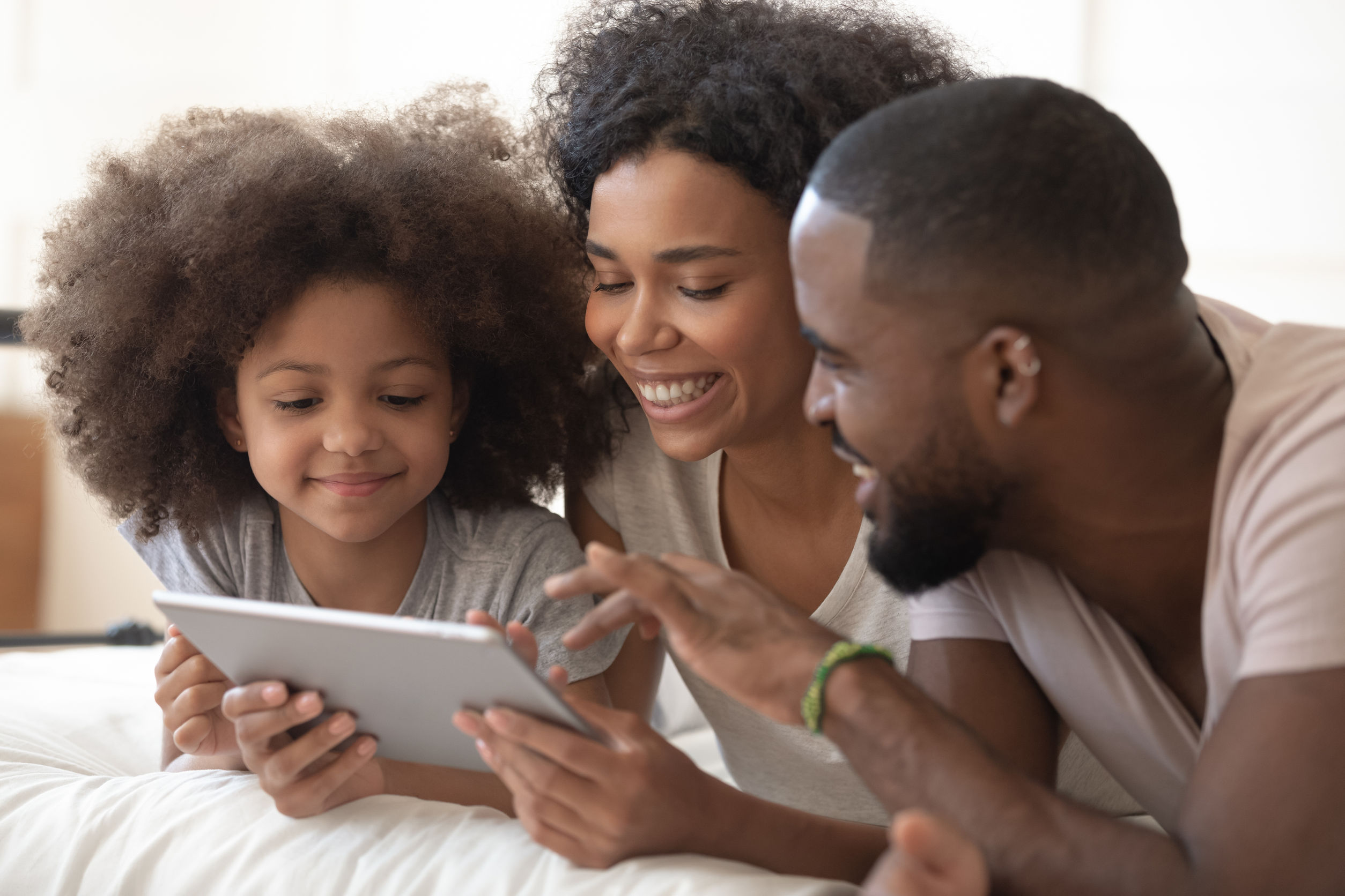 Happy black family of three lying on bed, using digital tablet together. Smiling african american girl with loving parents watching cartoons, photos or shopping, making order, ecommerce concept.