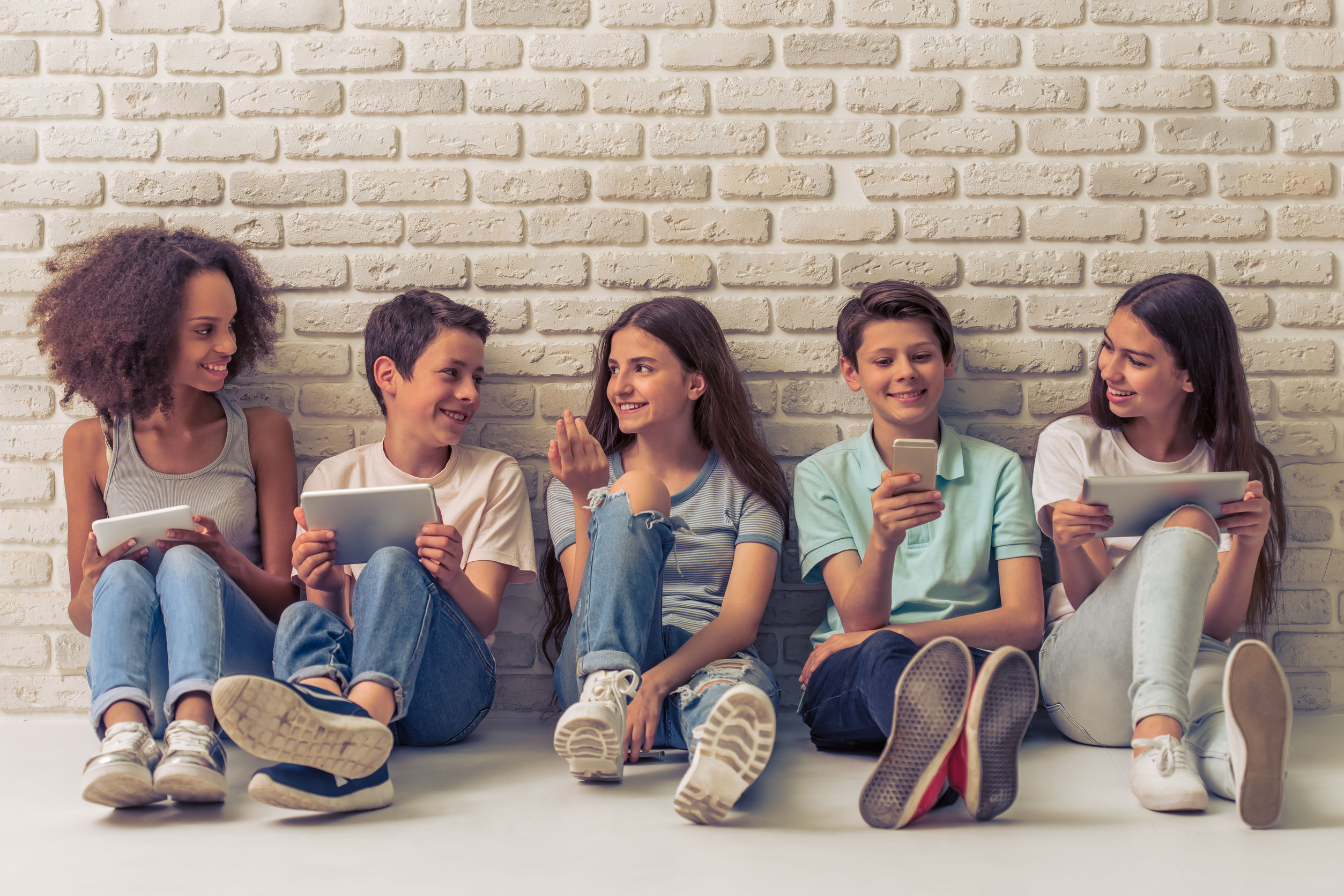 Group of teenage boys and girls is using gadgets, talking and smiling, sitting against white brick wall